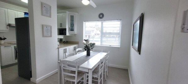 Une cuisine avec des meubles blancs, un réfrigérateur, une table à manger avec des chaises, une fenêtre avec des stores et une horloge au mur.