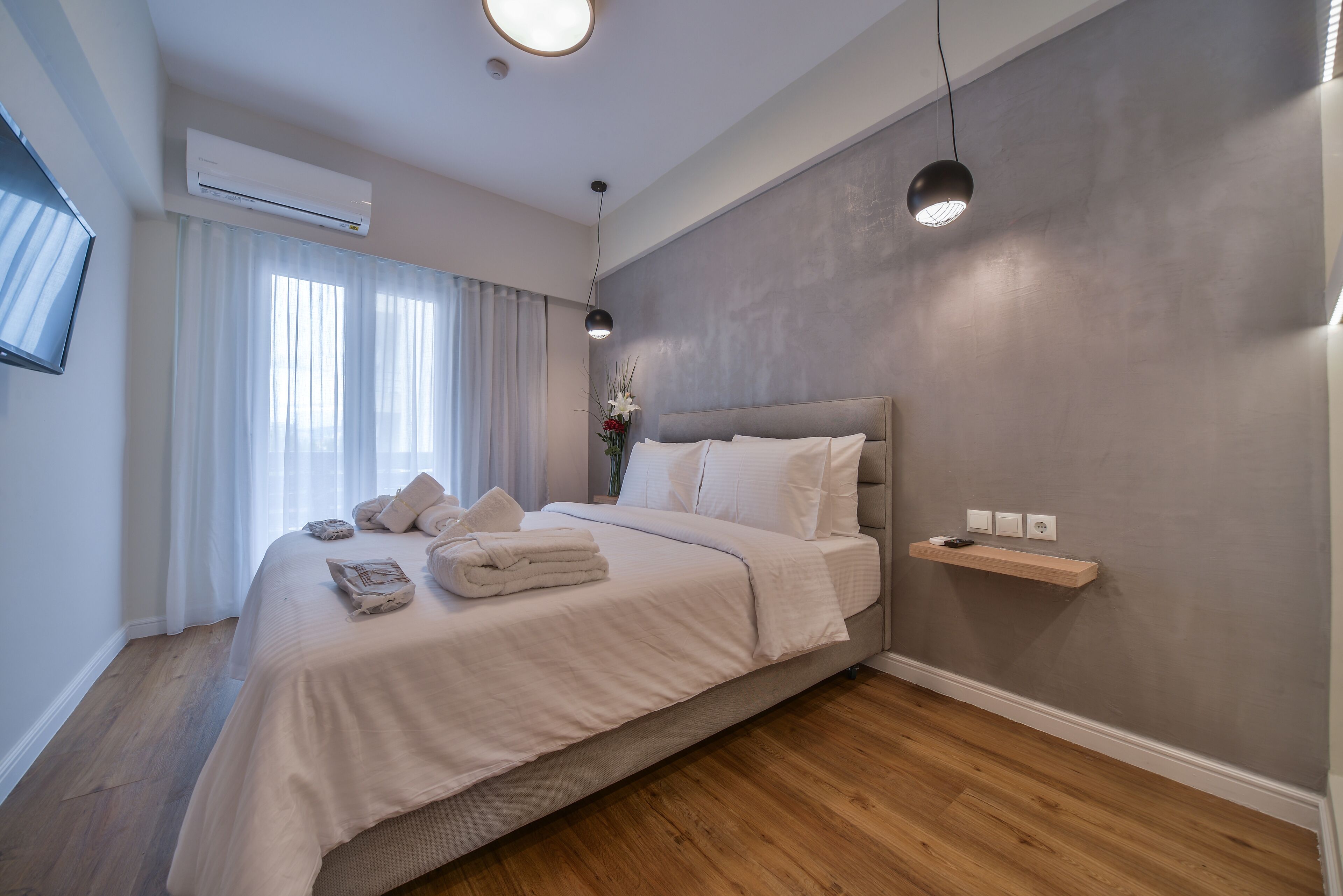 A modern bedroom with a large bed, a bedside table, a wall-mounted shelf, and a ceiling-mounted air conditioner.