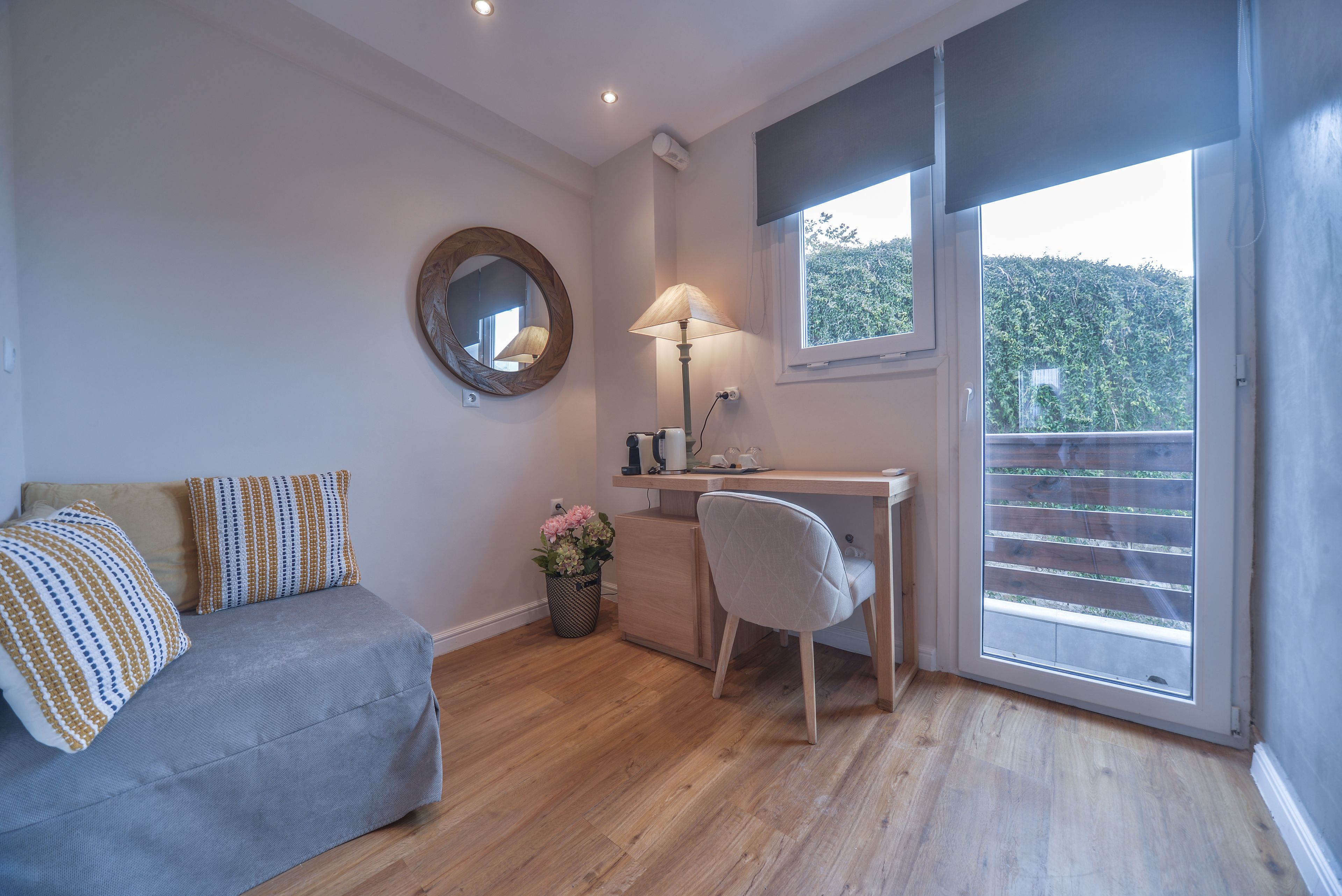 A room with a grey sofa, a desk with a lamp, a mirror, and a window with a view of greenery.