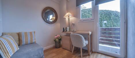 A room with a grey sofa, a desk with a lamp, a mirror, and a window with a view of greenery.