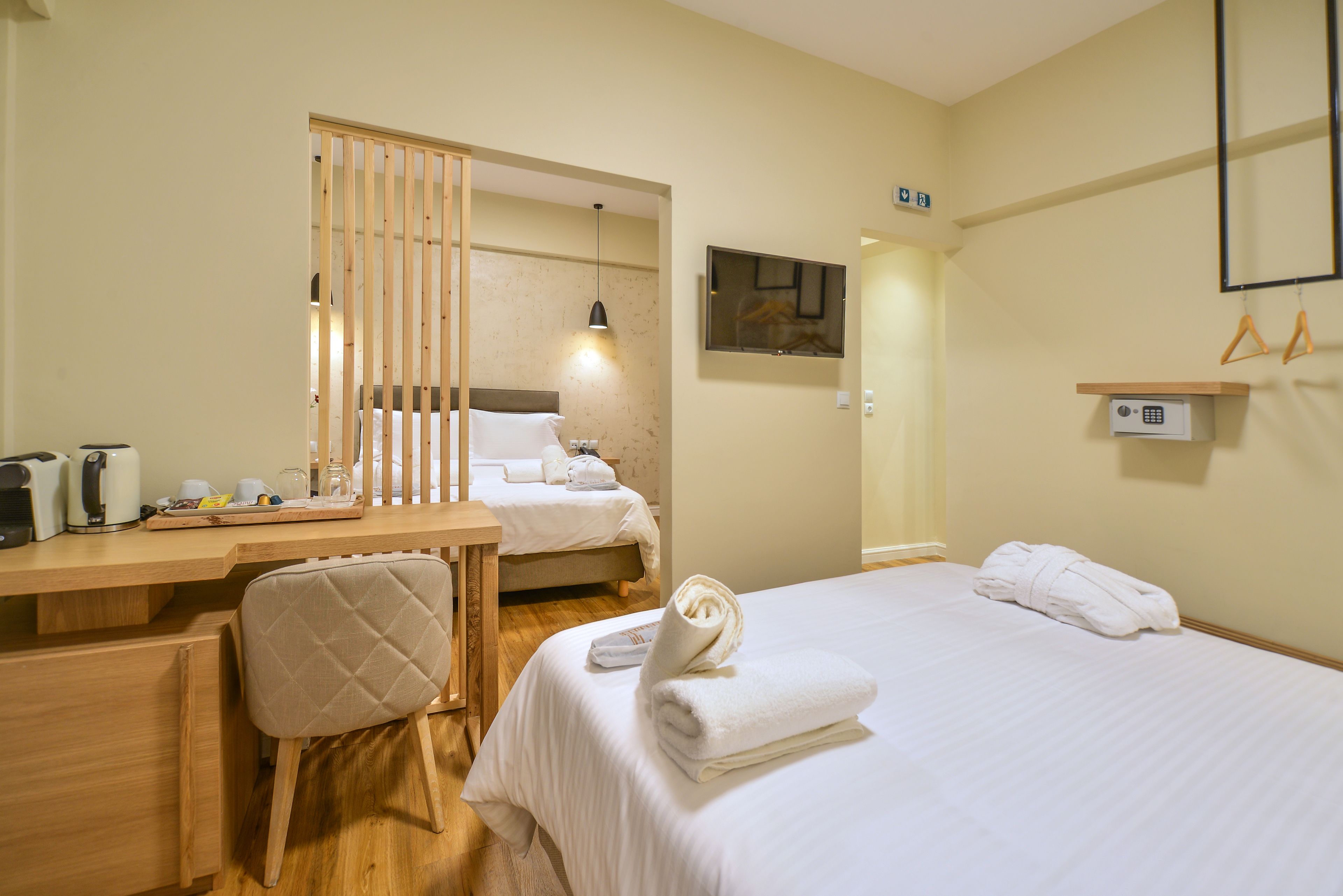 A hotel room with a bed, a desk, a chair, a bedside table, a mirror, and a small shelf with towels.
