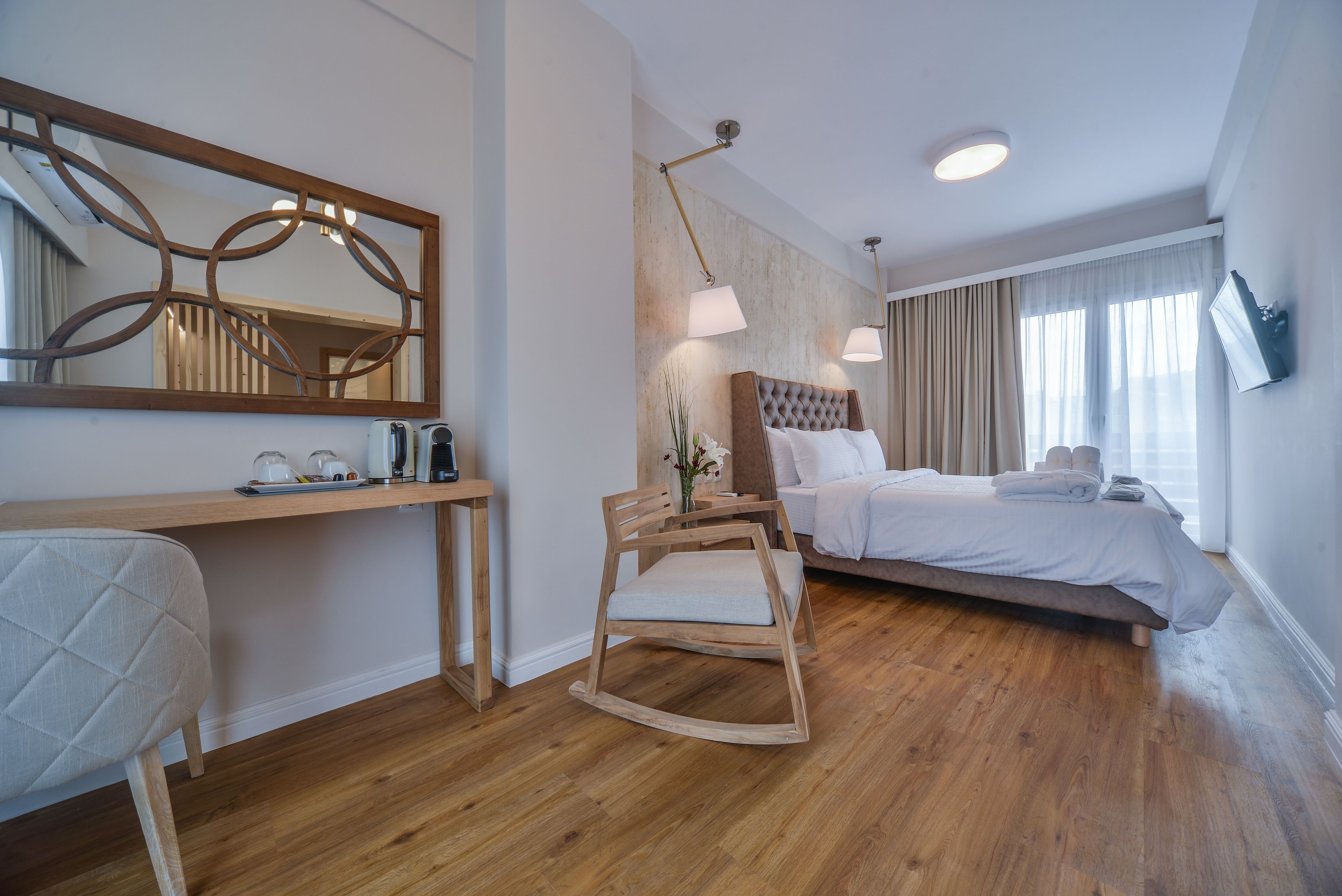 A modern hotel room with a wooden floor, a large bed with white linens, a wooden rocking chair, a desk with a chair, a large mirror, and a flat-screen TV mounted on the wall.