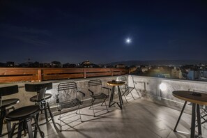 An outdoor terrace with metal chairs and tables, a wooden fence, and a cityscape view at night. - A.P. Acropolis View Apartments (Athens)