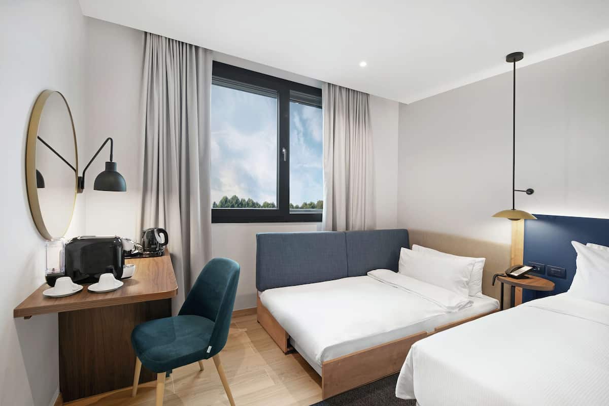 Una habitación de hotel moderna con una cama grande, un escritorio con una silla, una mesita con una lámpara y un ventanal con cortinas.
