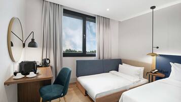 A modern hotel room with a large bed, a desk with a chair, a small table with a lamp, and a large window with curtains.