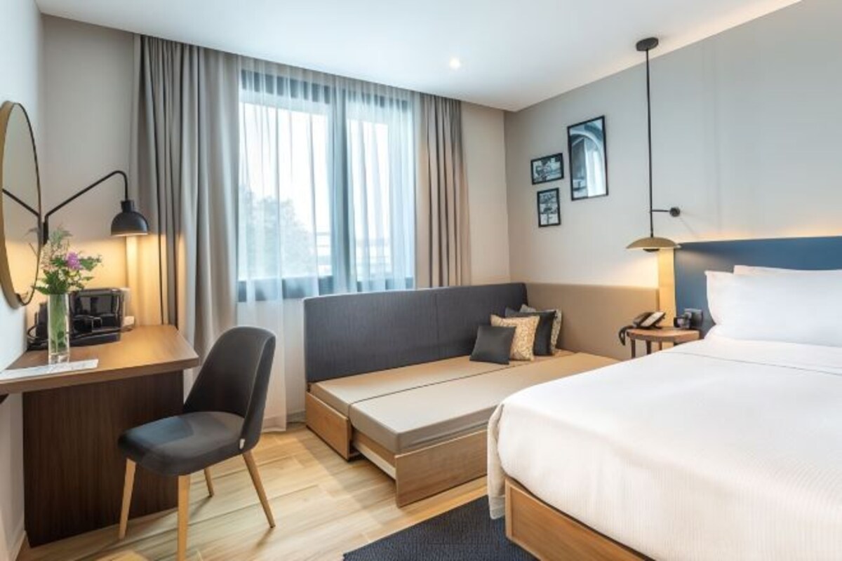 A modern hotel room with a bed, a desk, a chair, and a window with curtains.