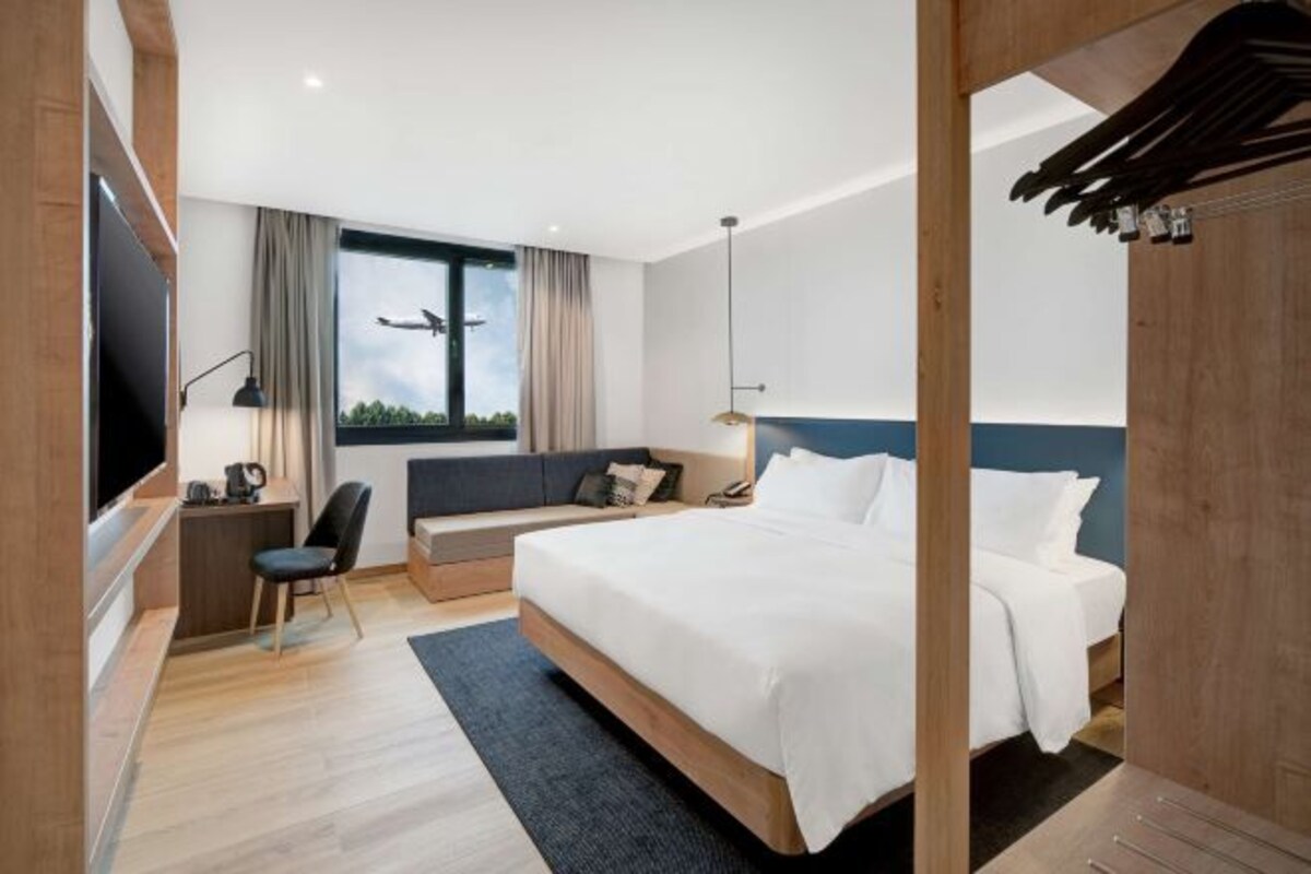 A modern hotel room with a large bed, a desk, a chair, a TV, and a window with a view of the sky.