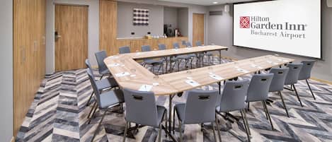 Un salón de conferencias con una larga mesa, sillas grises, una pantalla de proyector y piso alfombrado.