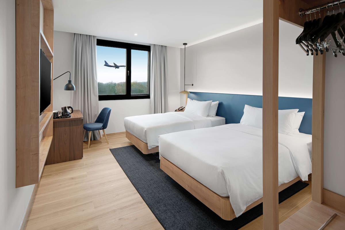 Habitación de hotel con dos camas, un escritorio, una silla, un televisor y una ventana con vista a un avión.