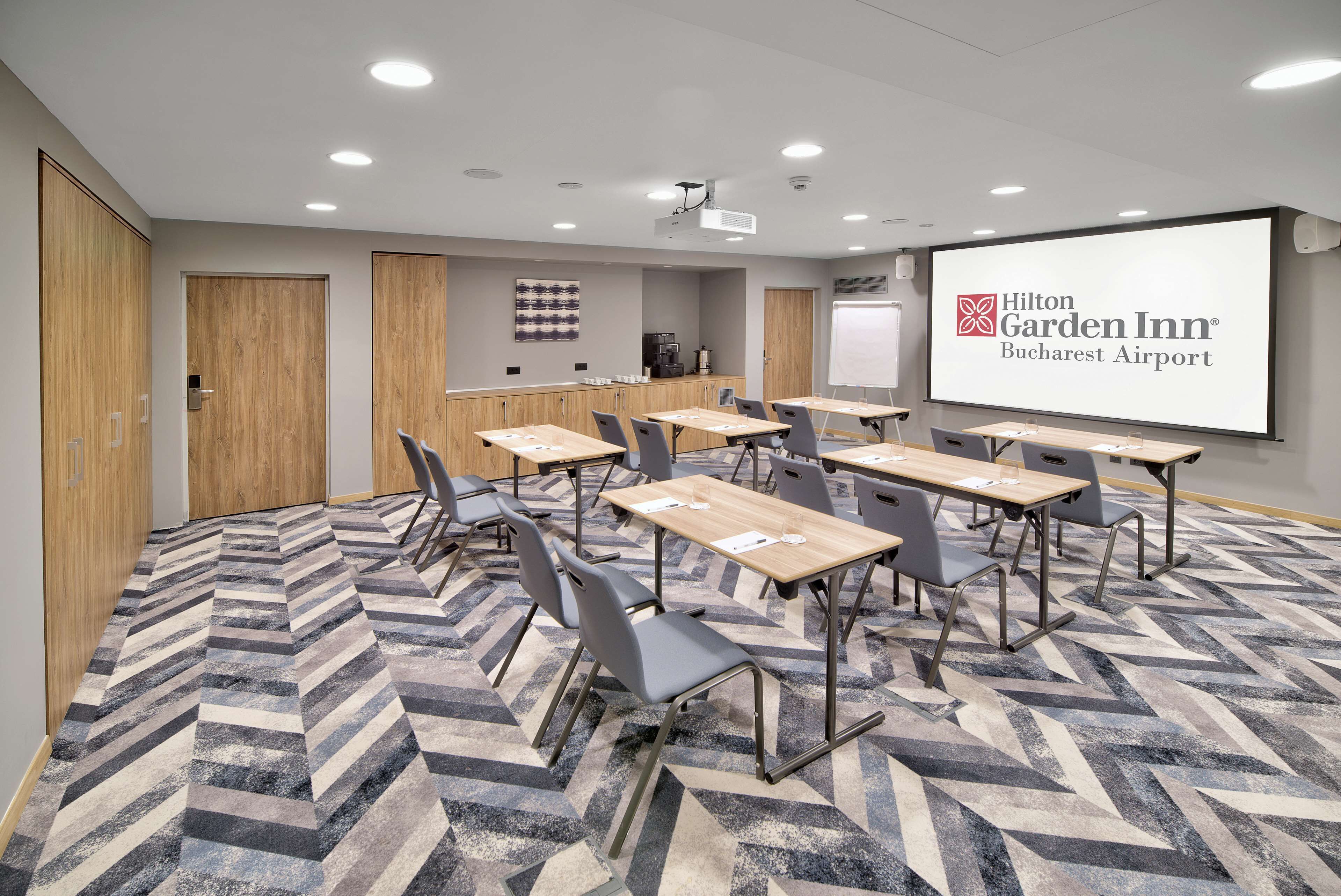 A conference room with a large screen displaying the Hilton Garden Inn Bucharest Airport logo, a projector, and a coffee station.