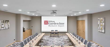 Ett konferensrum med ett långt bord, stolar, en projektorduk som visar Hilton Garden Inn Bucharest Airports logotyp och ett golv täckt av matta.