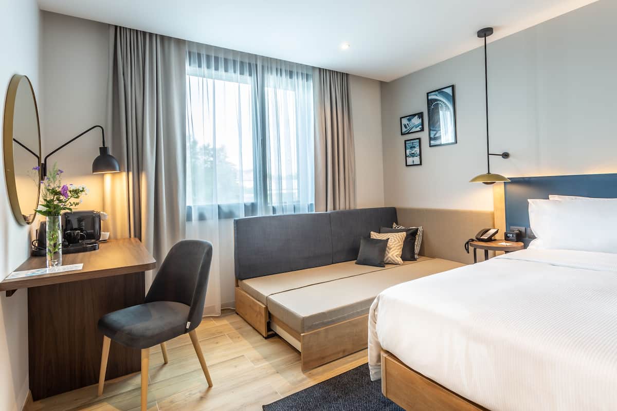 Habitación de hotel con cama, escritorio con lámpara, una silla, una mesita y ventana con cortinas.