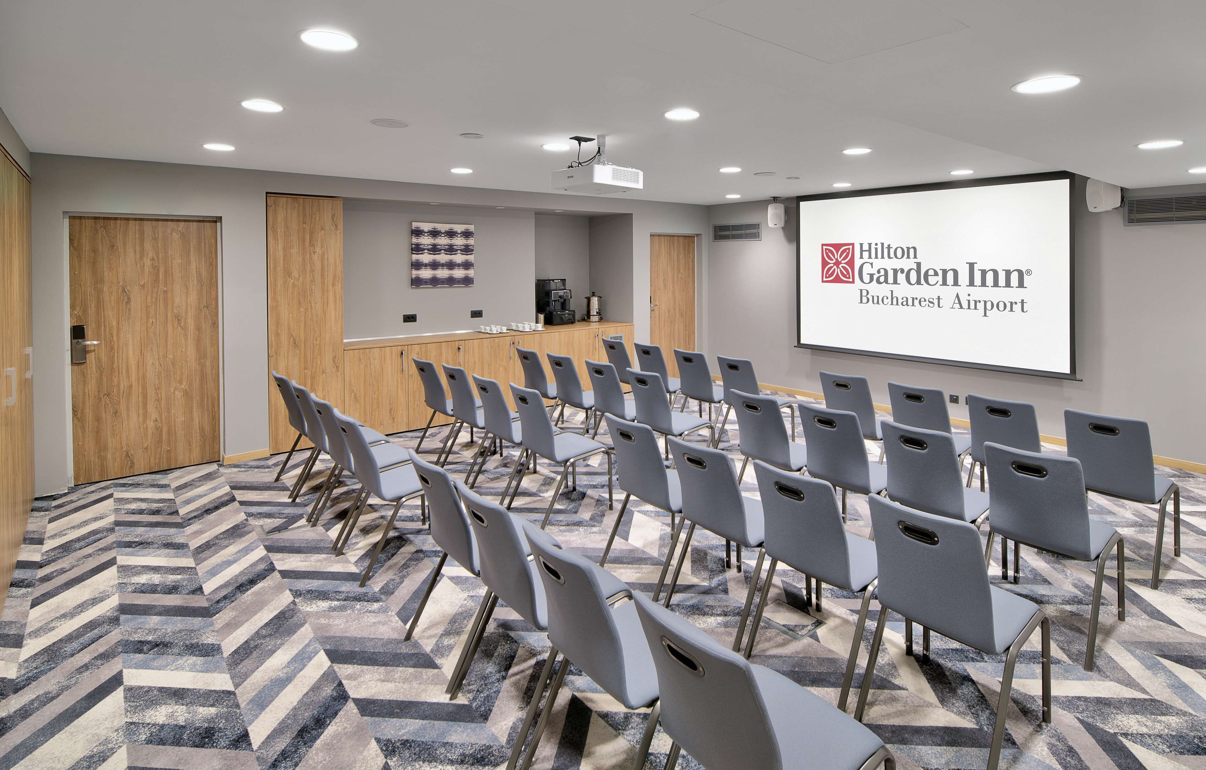 A conference room with rows of chairs, a podium, a projector screen displaying the Hilton Garden Inn Bucharest Airport logo, and a coffee station.