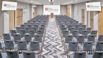A conference room with rows of grey chairs, a large screen displaying the Hilton Garden Inn Bucharest Airport logo, and a herringbone-patterned carpet.