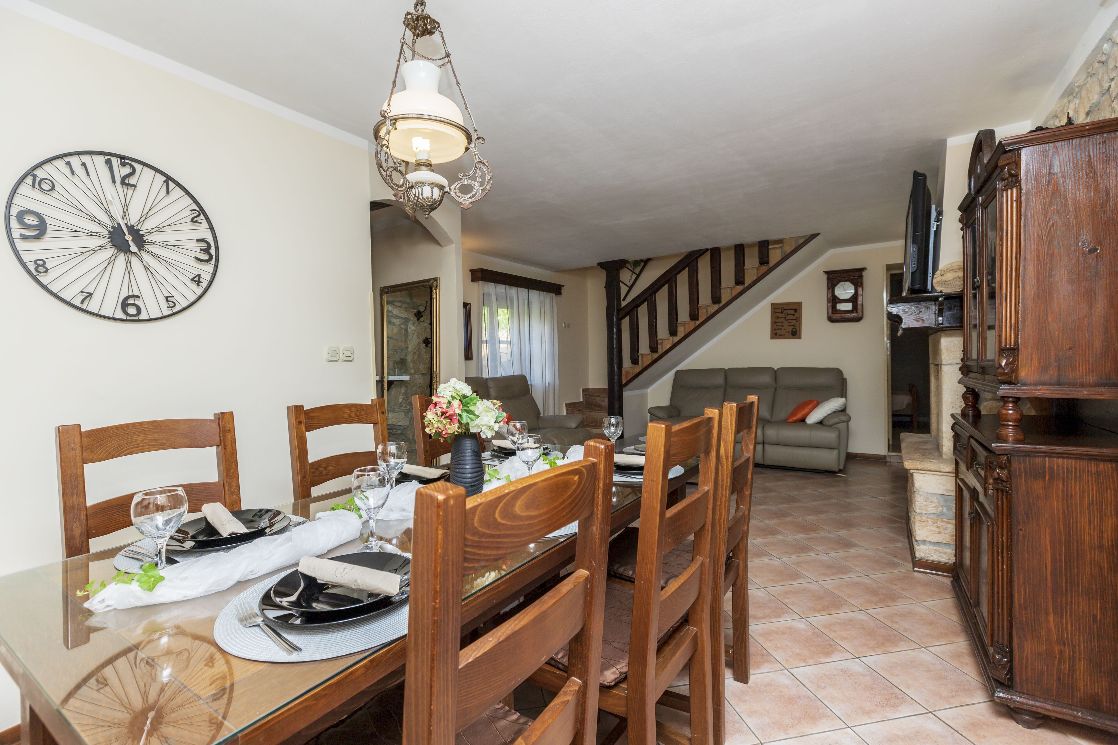 A dining area with a wooden table set for four, a large wall clock, and a staircase leading to another level.