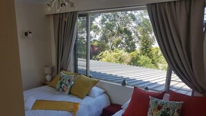 Une chambre d’hôtel avec deux lits, une grande fenêtre avec des rideaux, une table de chevet avec une lampe, et une vue sur la verdure à l’extérieur.