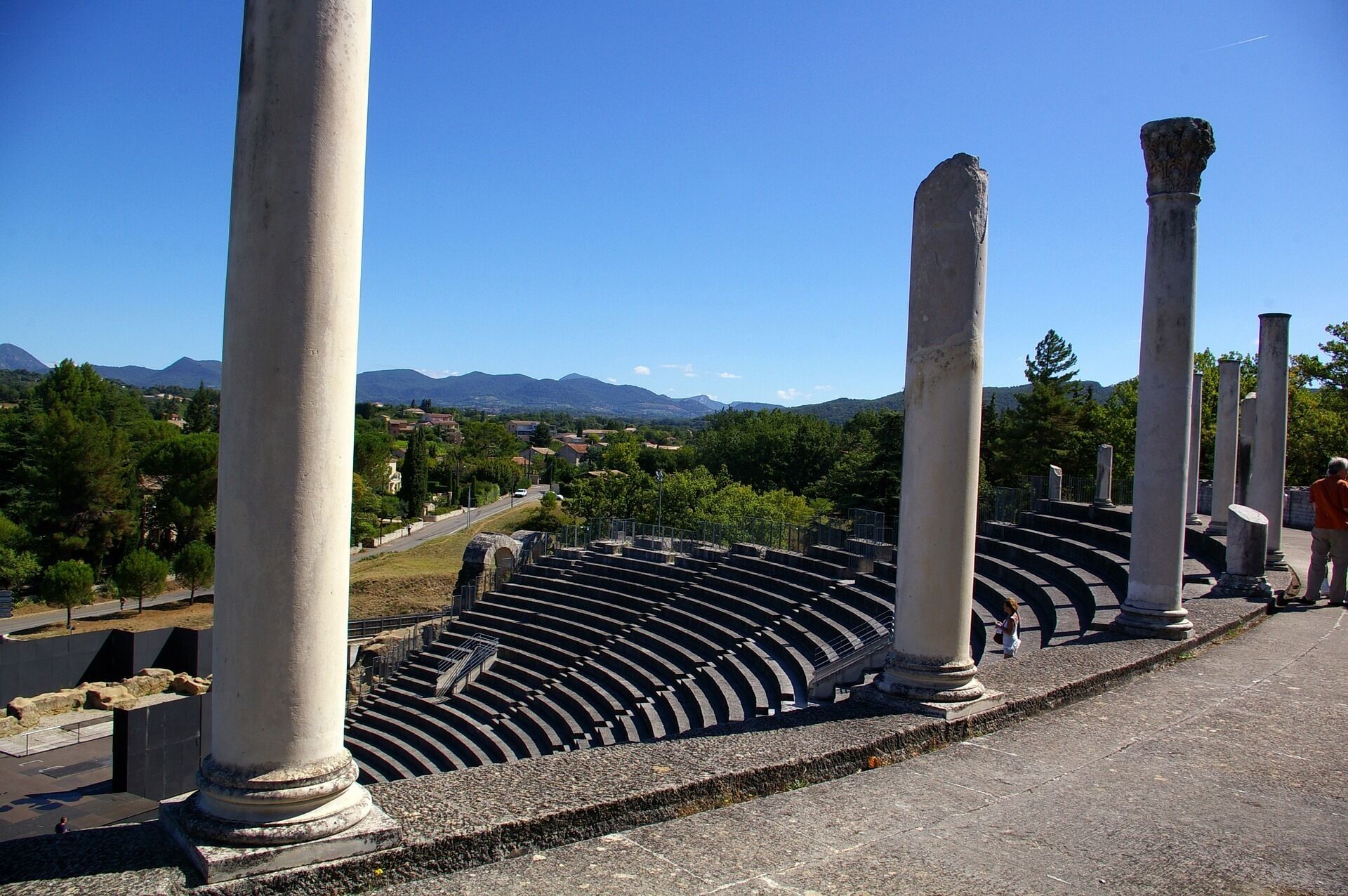 Ein uraltes Amphitheater mit Steinsäulen und terrassenförmigen Sitzreihen, umgeben von Grünpflanzen und Bergen im Hintergrund.