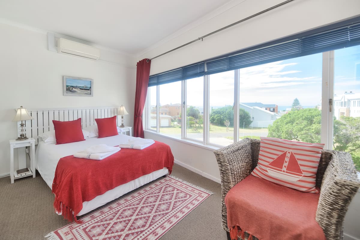 A bedroom with a large bed, a view of the outside, a window with curtains, and a wicker chair with a red cushion.