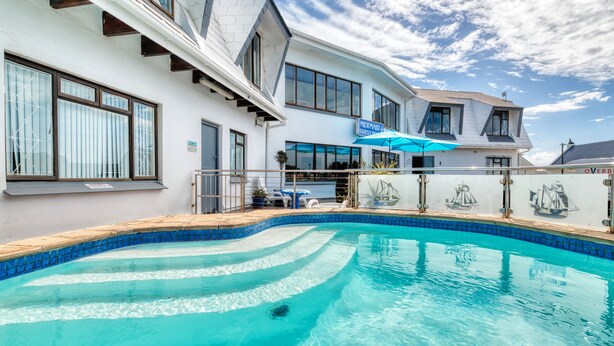 Ein Schwimmbecken mit Fliesenrand, ein Gebäude mit der Aufschrift "MERMAID GUESTHOUSE" und ein klarer, blauer Himmel.