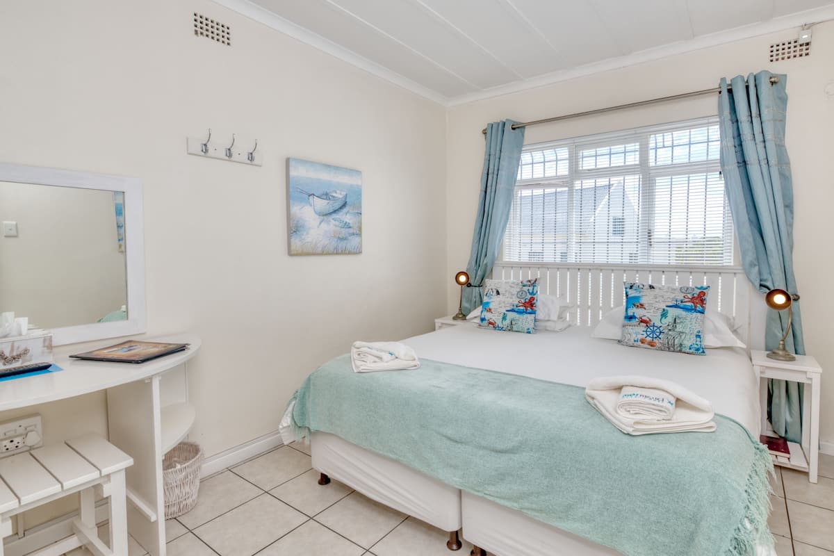 A bedroom with a bed, a desk, a mirror, and a window with curtains.