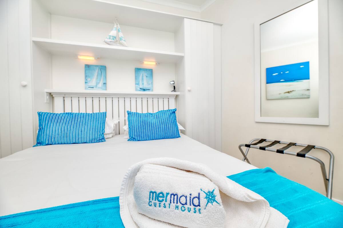 A hotel room with a bed, a towel with 'mermaid guest house' branding, and a painting of a sailboat on the wall.