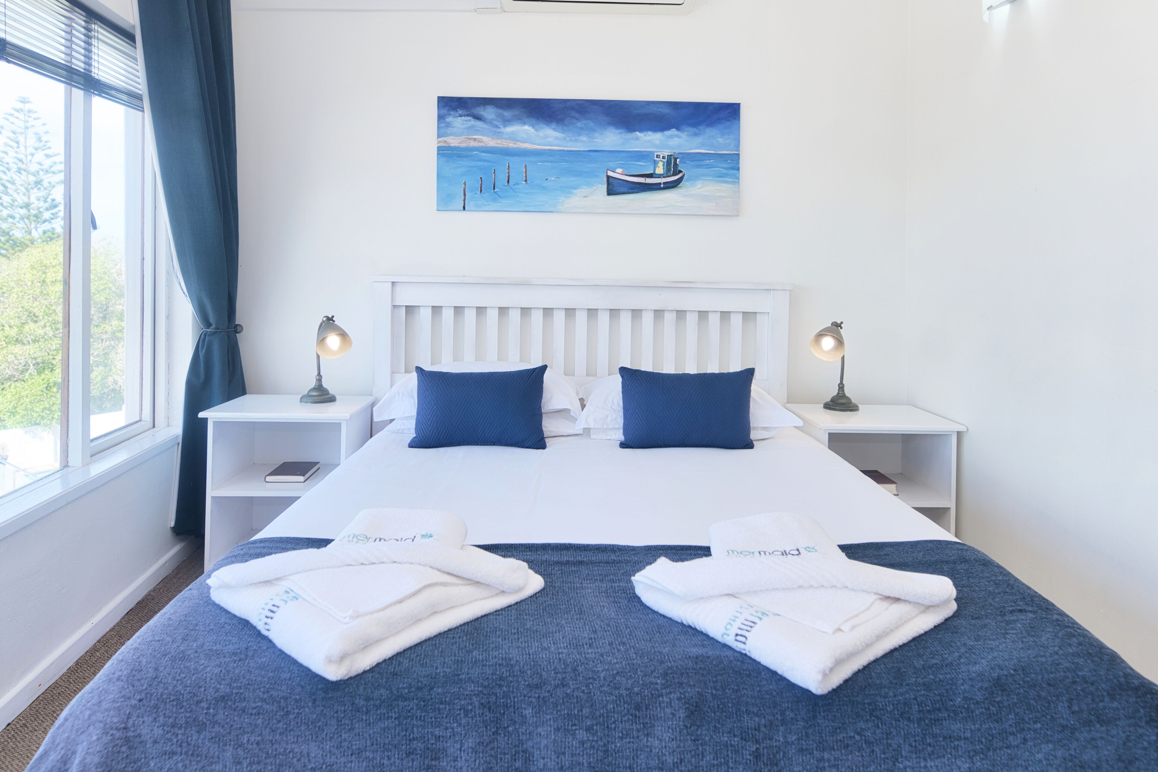 A hotel room with a large bed, two bedside tables, a painting of a boat on the wall, and a window with curtains.