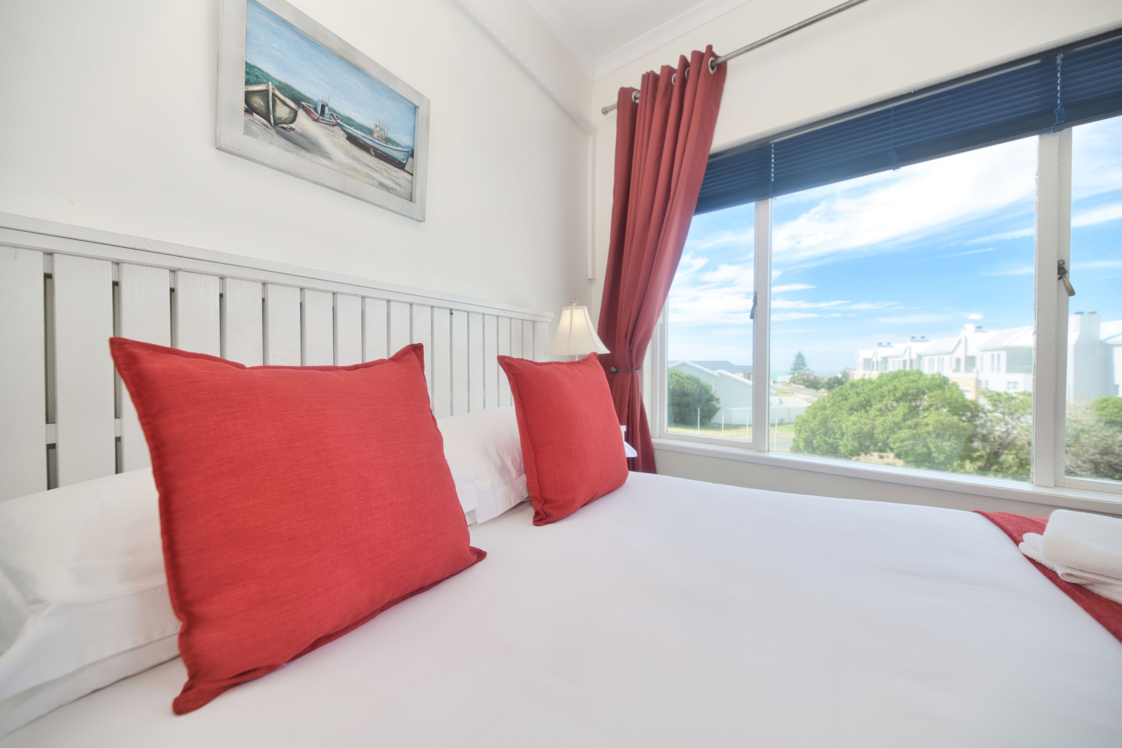 A hotel room with a large bed, red pillows, a white headboard, a window with a view of buildings and trees, and a painting on the wall.