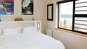 A hotel room with a bed, two posters on the wall, a window with a view, and a bedside table with a bottle. - Kite Mansion Penthouse (Langebaan)