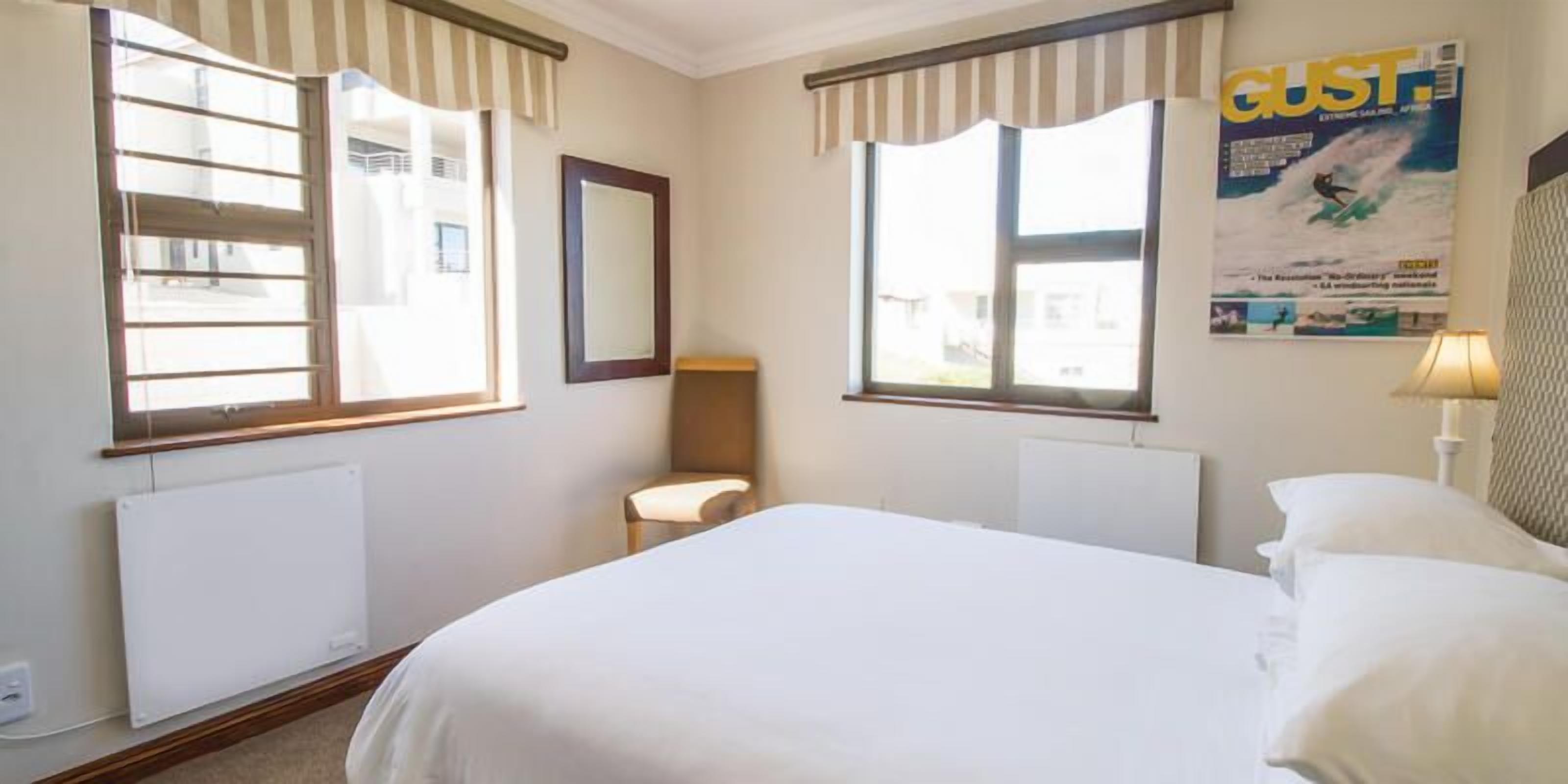 Een hotelkamer met een groot bed, twee ramen met gestreepte gordijnen, een radiator, een klein nachtkastje met een lamp en een poster aan de muur.