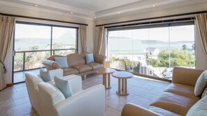 A spacious living area with a beige sofa set, a white sofa, and a small round table. Large windows provide a view of the sea and buildings. - Kite Mansion Penthouse (Langebaan)