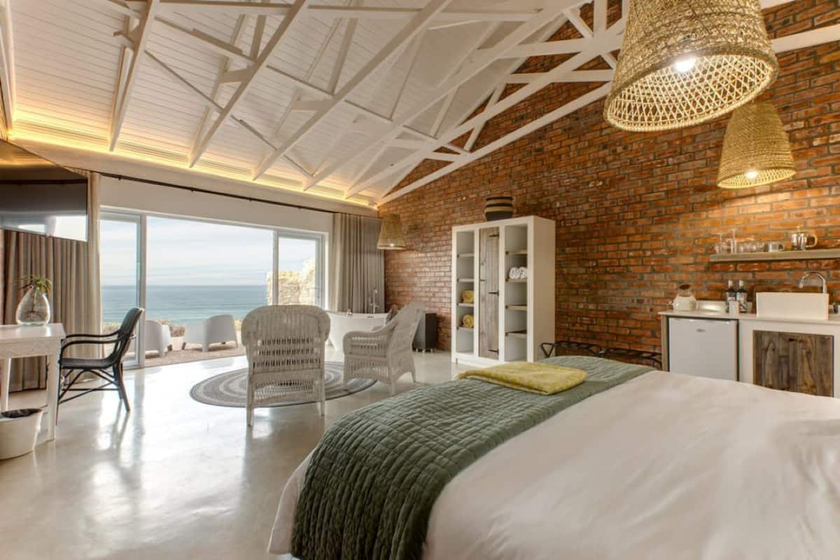 A modern bedroom with a large bed, a view of the sea, a brick wall, and a white ceiling structure.