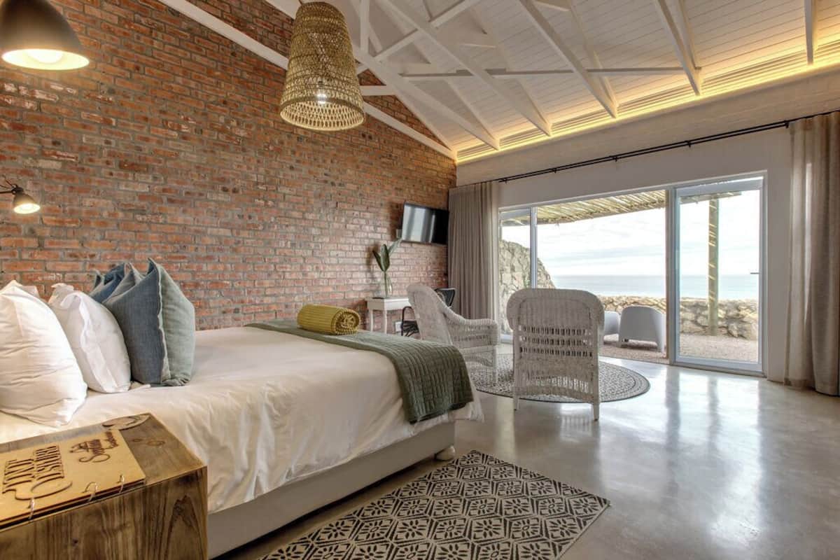 A modern bedroom with a brick wall, a large bed, a TV, and a view of the beach through a sliding glass door.