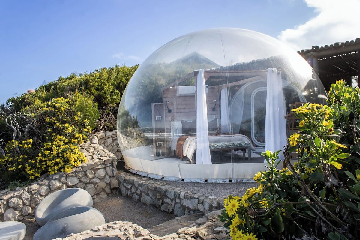 A transparent dome structure with a bed inside, surrounded by stone walls and greenery.