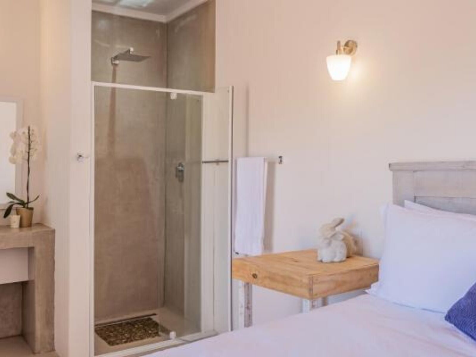 Una camera da letto con un letto, un comodino in legno, un tavolino, una doccia e un punto luce a parete.