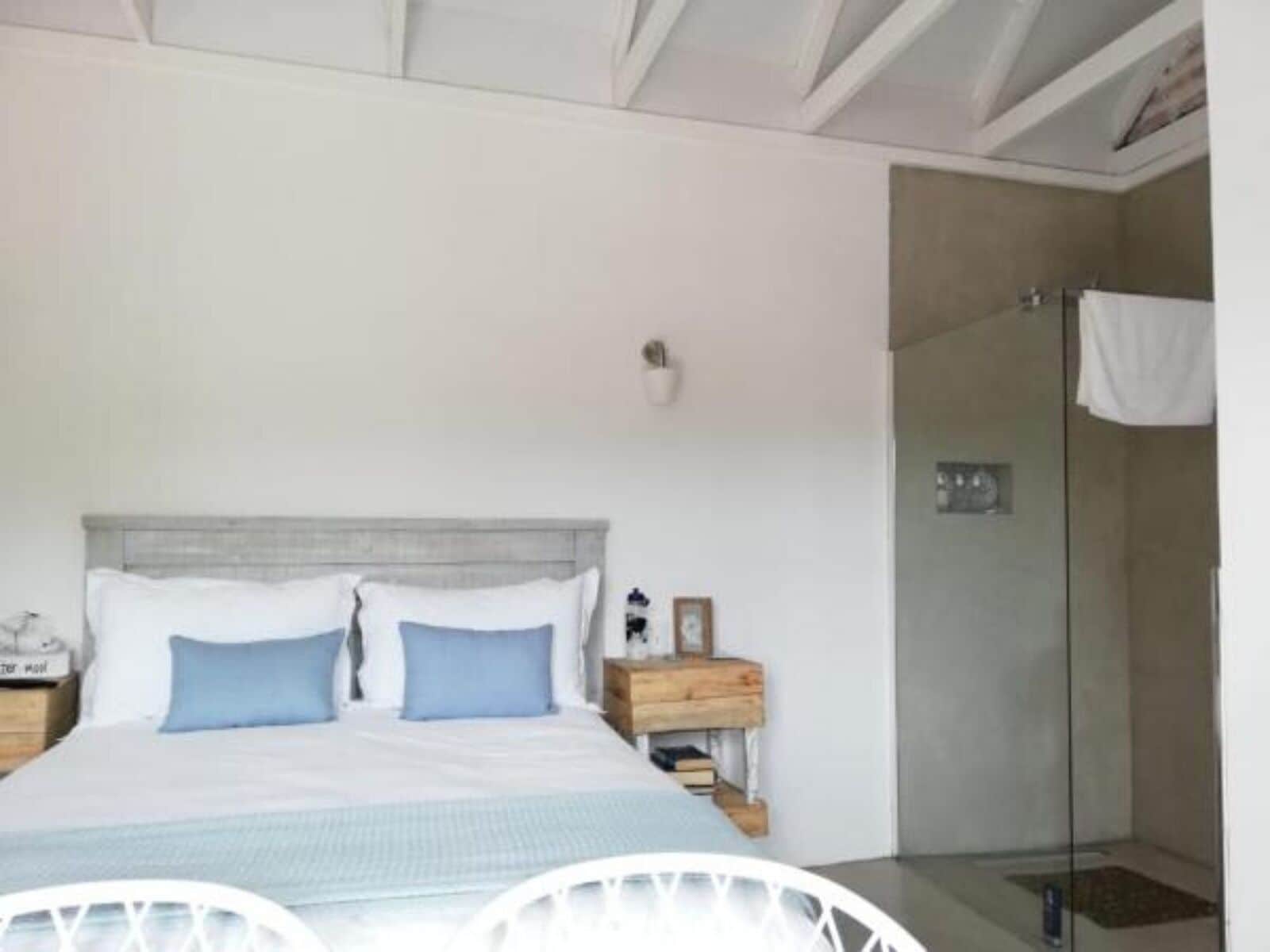 Un dormitorio con cama, mesitas de noche, cómoda de madera, una silla blanca y cabina de ducha con paredes de vidrio.