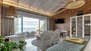 Un dormitorio moderno con una cama grande, una bañera, sillas de mimbre y vistas al océano.
