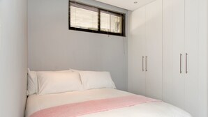 A bedroom with a bed, white walls, a window with blinds, and a wardrobe with two doors. - Wild Olive 11 (Windhoek)