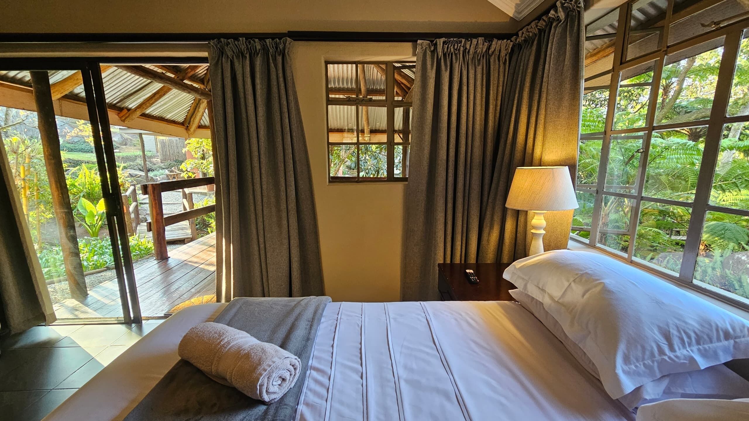 Habitación con cama, mesita de noche, lámpara y vistas al verdor a través de amplios ventanales.
