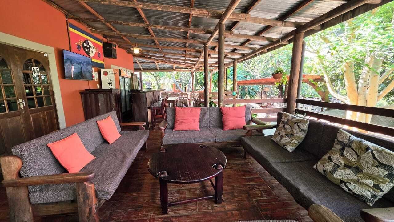 A covered outdoor seating area with wooden furniture, a television, and a view of greenery.