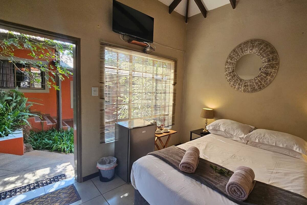 A hotel room with a bed, a television, a window with blinds, a small table, and a trash bin.