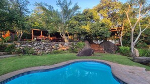 Una piscina con borde de piedra, rodeada de césped, árboles y una estructura de madera con techo.