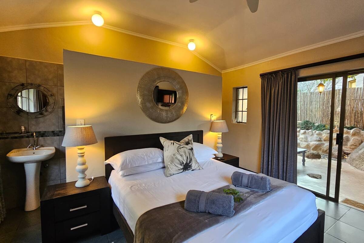 A hotel room with a bed, two bedside lamps, a round mirror, a window with curtains, and a view of an outdoor area.