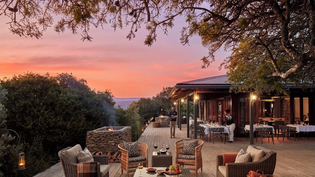 An outdoor dining area with wicker furniture, a stone fireplace, and a view of the ocean at sunset. - Ecca Lodge (Makhanda)