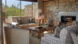 A cozy living room with a stone wall, large windows, and a fireplace. - Ecca Lodge (Makhanda)