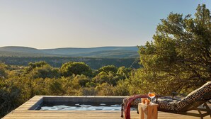 A wooden deck with a hot tub, a lounge chair, and a table with drinks and snacks. - Ecca Lodge (Makhanda)