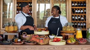 Two waitresses serving food and drinks in a restaurant with a wine cellar in the background. - Ecca Lodge (Makhanda)