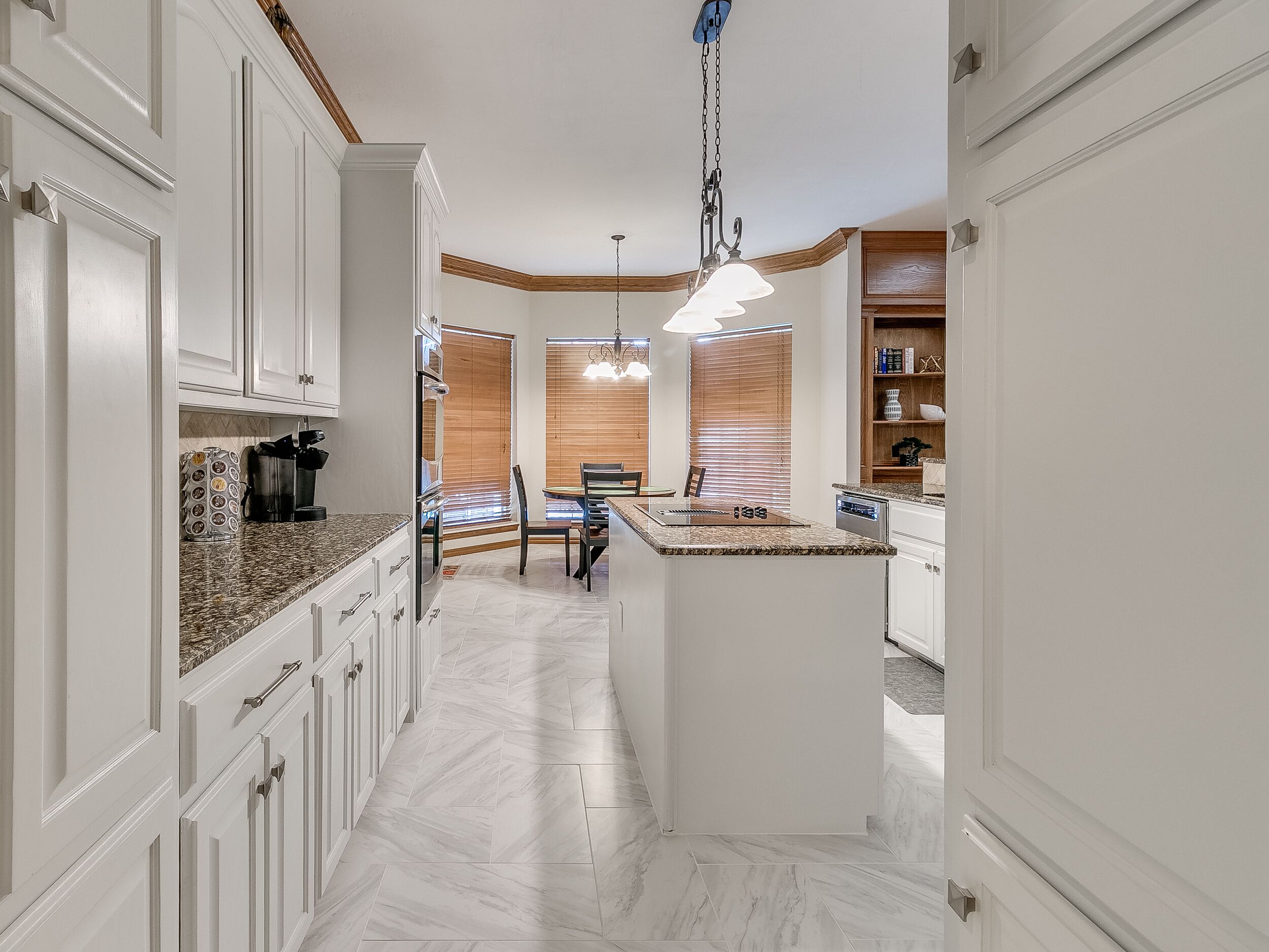 A modern kitchen with white cabinetry, granite countertops, a central island, and a dining area with wooden blinds.