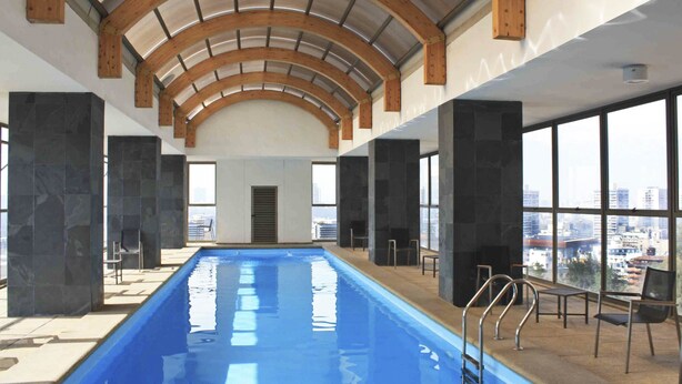 An indoor swimming pool with a high arched wooden ceiling, stone pillars, and large windows offering a city view.