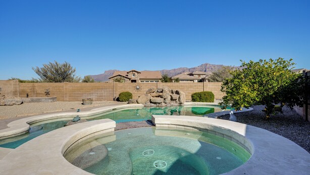 A swimming pool with a hot tub, surrounded by a landscaped area with a view of mountains and a house in the background.