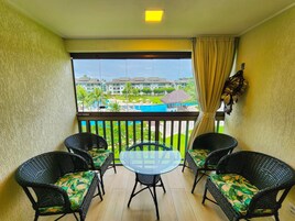 A balcony with wicker chairs and a glass table, overlooking a pool and resort buildings.